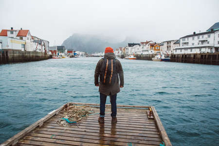 Traveler man with a backpack wearing a red hat standing on the background of fishing houses, ships, mountain and lake wooden pier. Travel lifestyle concept. Shoot from the backの写真素材