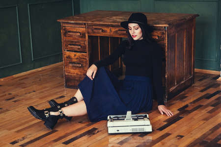 Beautiful woman in a hat sits near oak table with vintage typewriterの写真素材