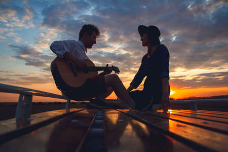 Silhouette of a man with a guitar and a woman in a hat sitting on the roof of a car on the background of the sunset. Silhouette romance concept.の写真素材