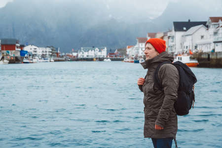 Traveler man with a backpack wearing a red hat standing on the background of fishing houses, ships, mountain and lake wooden pier. Travel lifestyle concept.の写真素材