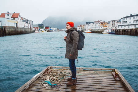 Traveler man with a backpack wearing a red hat standing on the background of fishing houses, ships, mountain and lake wooden pier. Travel lifestyle concept.の写真素材