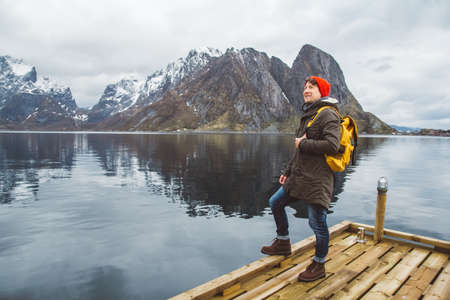 Traveler man with a yellow backpack wearing a red hat standing on the background of mountain and lake wooden pier. Travel lifestyle conceptの写真素材