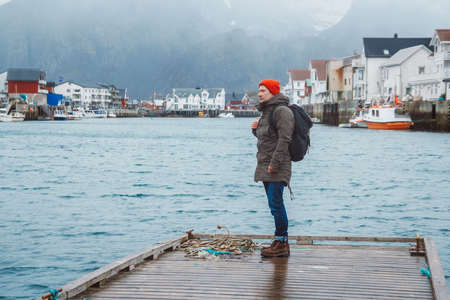 Traveler man with a backpack wearing a red hat standing on the background of fishing houses, ships, mountain and lake wooden pier. Travel lifestyle concept.の写真素材