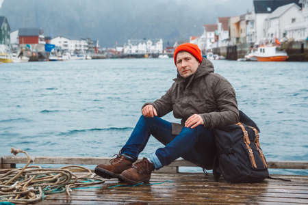 Traveler man with a backpack wearing a red hat sits on the background of fishing houses, ships, mountain and lake wooden pier. Travel lifestyle concept.の写真素材