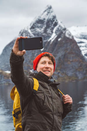 Portrait traveler man taking self-portrait a photo with a smartphone. Tourist in a yellow backpack standing on a background of a mountain. Traveler walks, takes photo in the mountains.の写真素材