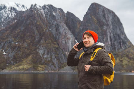 Portrait traveler man talking on mobile phone. Tourist in a yellow backpack standing on a background of a mountain and a lake. Traveler walks, smiling man calling friends. Telephone conversation.の写真素材