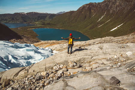 Traveler man with a yellow backpack wearing a red hat sitting on the shore on the background of mountain and lake. Space for your text message or promotional content. Travel lifestyle concept. Shoot from the back.の写真素材