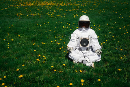 Futuristic astronaut in a helmet sits on a green lawn among flowersin a meditative position.の写真素材