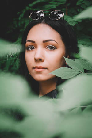 Portrait of a beautiful girl in black clothes and glasses from the sun against the background of green leaves. A look at the cameraの写真素材