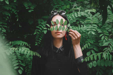 Beautiful girl in black clothes and glasses from the sun holds a twig in her hand near her face against the background of green leavesの写真素材