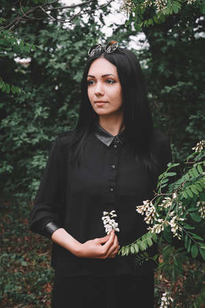 Beautiful girl in black clothes and glasses from the sun holds a white flower in her hand against the background of green leavesの写真素材