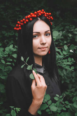 Beautiful girl in black dress and red decorations that holds a twig in her hand against the background of the forestの写真素材