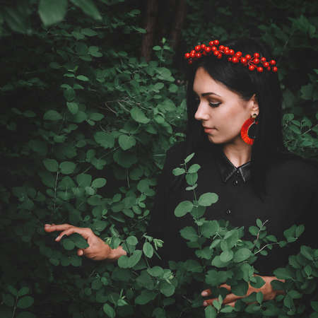 Beautiful girl in black dress and red decorations that holds a twig in her hand against the background of the forest. Space for your text message or promotional contentの写真素材