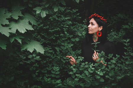 Beautiful girl in black dress and red decorations that holds a twig in her hand against the background of the forest. Space for your text message or promotional contentの写真素材