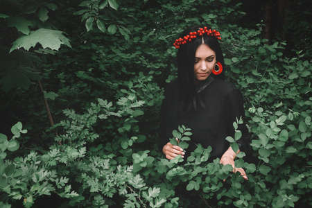 Beautiful girl in black dress and red decorations that holds a twig in her hand against the background of the forest. Space for your text message or promotional contentの写真素材