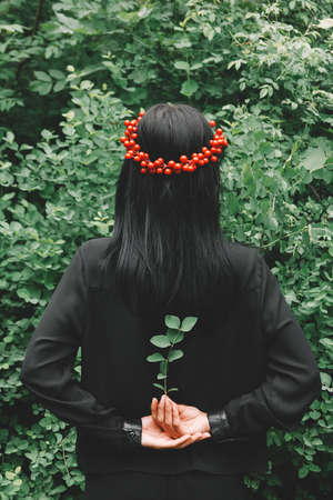 Beautiful girl with black hair and red accessories that holds a twig in her arms behind her back against the background of the forestの写真素材