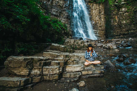 Beautiful girl with red hair in a hat and shirt meditating on rocks in a lotus position against a waterfall. Space for your text message or promotional contentの写真素材