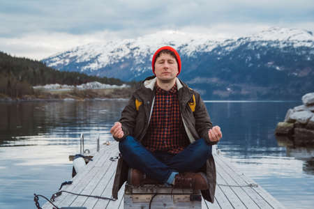 Traveler man in a meditative position sitting on a wooden pier on the background of a mountain and a lake. Space for your text message or promotional content.の写真素材