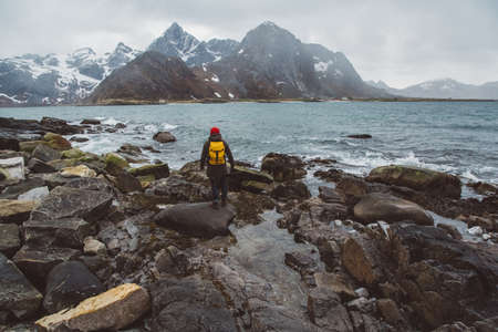 Traveler man with a yellow backpack wearing a red hat standing on the background of mountains and sea. Travel lifestyle concept. Shoot from the backの写真素材