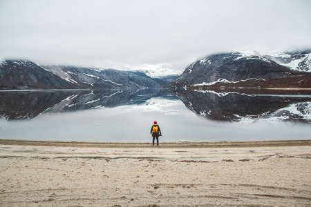 Traveler man with a yellow backpack wearing a red hat standing on the background of mountains and lake enjoying landscape. Travel lifestyle concept. Shoot from the backの写真素材