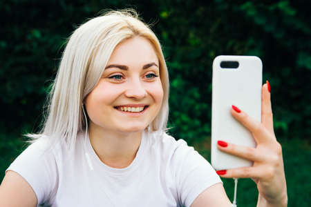 Portrait of beautiful blonde girl in headphones and smartphone in hands sitting on green background. She is talking on the phone through headphones. Lifestyle concept. Copy, empty space for textの写真素材