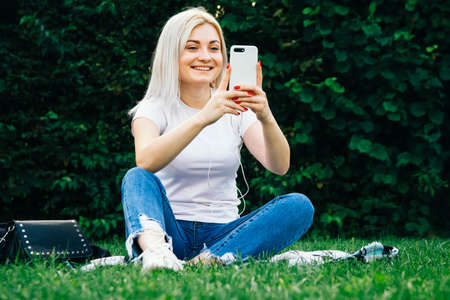 Beautiful blonde girl in headphones and smartphone in hands sits on green grass. She is talking on the phone through headphones. Lifestyle concept. Copy, empty space for textの写真素材