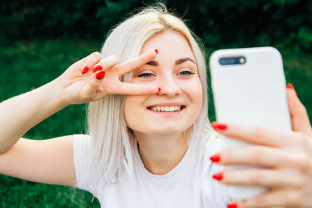 Portrait of beautiful blonde girl in headphones and smartphone in hands sitting on green background. She is talking on the phone through headphones. Time to take selfie. Copy, empty space for textの写真素材