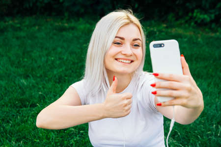 Portrait of beautiful blonde girl in headphones and smartphone in hands sitting on green background. She is talking on the phone through headphones. Time to take selfie. Copy, empty space for textの写真素材