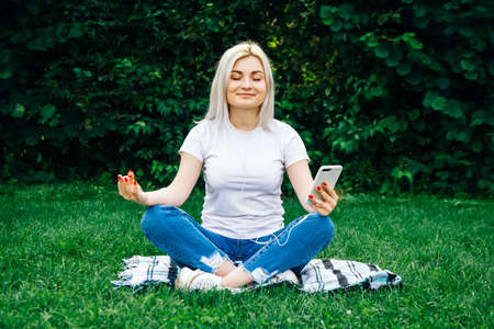 Beautiful blonde girl in headphones and smartphone in hands sits in meditative pose listening to music on green grass. Lifestyle concept. Copy, empty space for textの写真素材