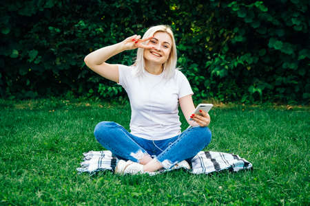 Beautiful blonde girl in headphones and smartphone in hands sits on green grass. Lifestyle concept. Copy, empty space for textの写真素材