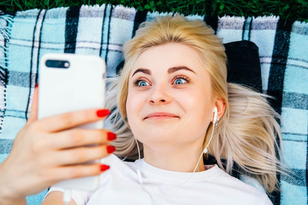 Portrait of beautiful blonde girl in headphones and smartphone in hands lying on rug green...