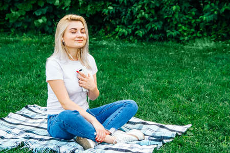 Beautiful blonde girl in headphones and smartphone in hands sits on green grass. Lifestyle concept. Copy, empty space for textの写真素材