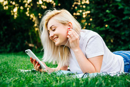 Beautiful blonde girl in headphones and smartphone in hands listens to music lying on green background. Lifestyle concept. Copy, empty space for textの写真素材