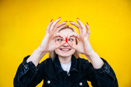 Crazy beautiful blonde girl in a black jacket smiles and simulates glasses with her hands against...