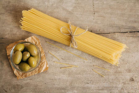 Long yellow raw spaghetti and green olives in glass bowl on wooden background. Top view. Copy, empty space for text.の写真素材