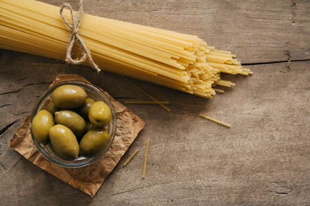 Long yellow raw spaghetti and green olives in glass bowl on wooden background. Top view. Copy, empty space for text.の写真素材