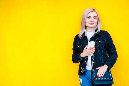 Beautiful blonde girl in headphones and smartphone in hands is talking on the phone through headphones on yellow wall background. Time to take selfie. Copy, empty space for text.の写真素材