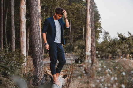 Handsome man posing outdoors in forest stands barefoot on a log, wearing jacket. Place for text or advertising.の写真素材