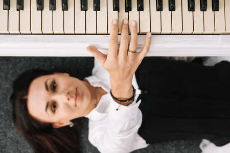 Beautiful woman dressed in a white dress with a black corset lies on the floor near white piano playing on the keys. Place for text or advertising. View from above.の写真素材