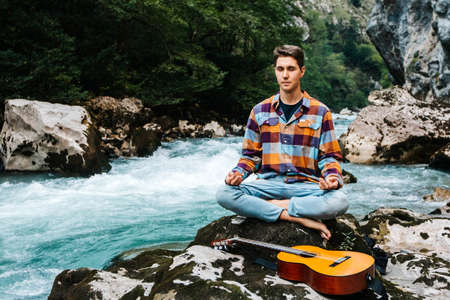 Man in a meditative position with guitar sitting on the bank of a mountain river on a background of rocks and forest. Concept of freedom relaxation. Place for text or advertising.の写真素材