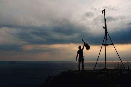 Silhouette of a man holding a guitar on a background of clouds and the sea at sunset. Shoot from the back. Concept of freedom relaxation. Place for text or advertising.の写真素材
