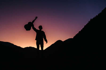 Silhouette of a man holding a guitar on a background of mountains and sunset. Concept of freedom relaxation. Place for text or advertising.の写真素材