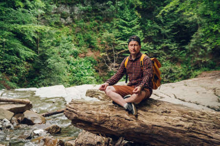 Traveler man in a meditative position with a backpack sitting on a tree trunk against the background of the forest and the river. Space for your text message or promotional contentの写真素材