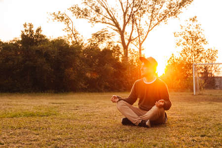 Young handsome man in a meditative position sits on a lawn on a background of forest and sunset. Concept of freedom relaxation. Place for text or advertising.の写真素材
