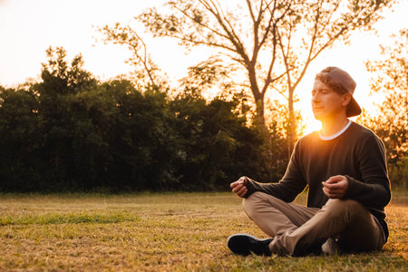 Young handsome man in a meditative position sits on a lawn on a background of forest and sunset. Concept of freedom relaxation. Place for text or advertising.の写真素材