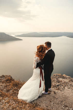 Amazing wedding couple, bride and groom holding hands on a mountains and river background. Cute girl in white dress, men in business suit. Mountains landscape and rivers in the sunset.の写真素材