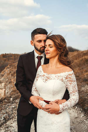 Amazing wedding couple, bride and groom holding hands on a mountains and river background. Cute girl in white dress, men in business suit. Mountains landscape and rivers in the sunset.の写真素材