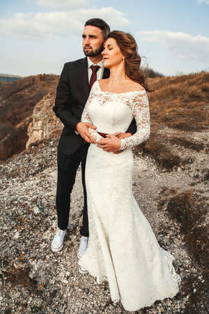 Amazing wedding couple, bride and groom holding hands on a mountains and river background. Cute girl in white dress, men in business suit. Mountains landscape and rivers in the sunset.の写真素材