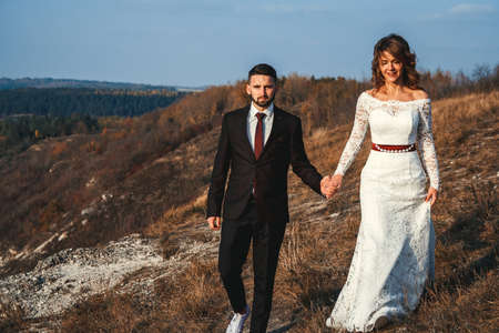 Amazing wedding couple, bride and groom holding hands on a mountains and river background. Cute girl in white dress, men in business suit. Mountains landscape and rivers in the sunset.の写真素材