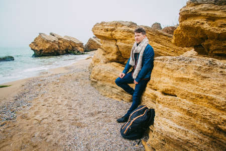 Traveler man with backpack standing on sandy beach in the middle of rocks against sea background. Place for text or advertising.の写真素材